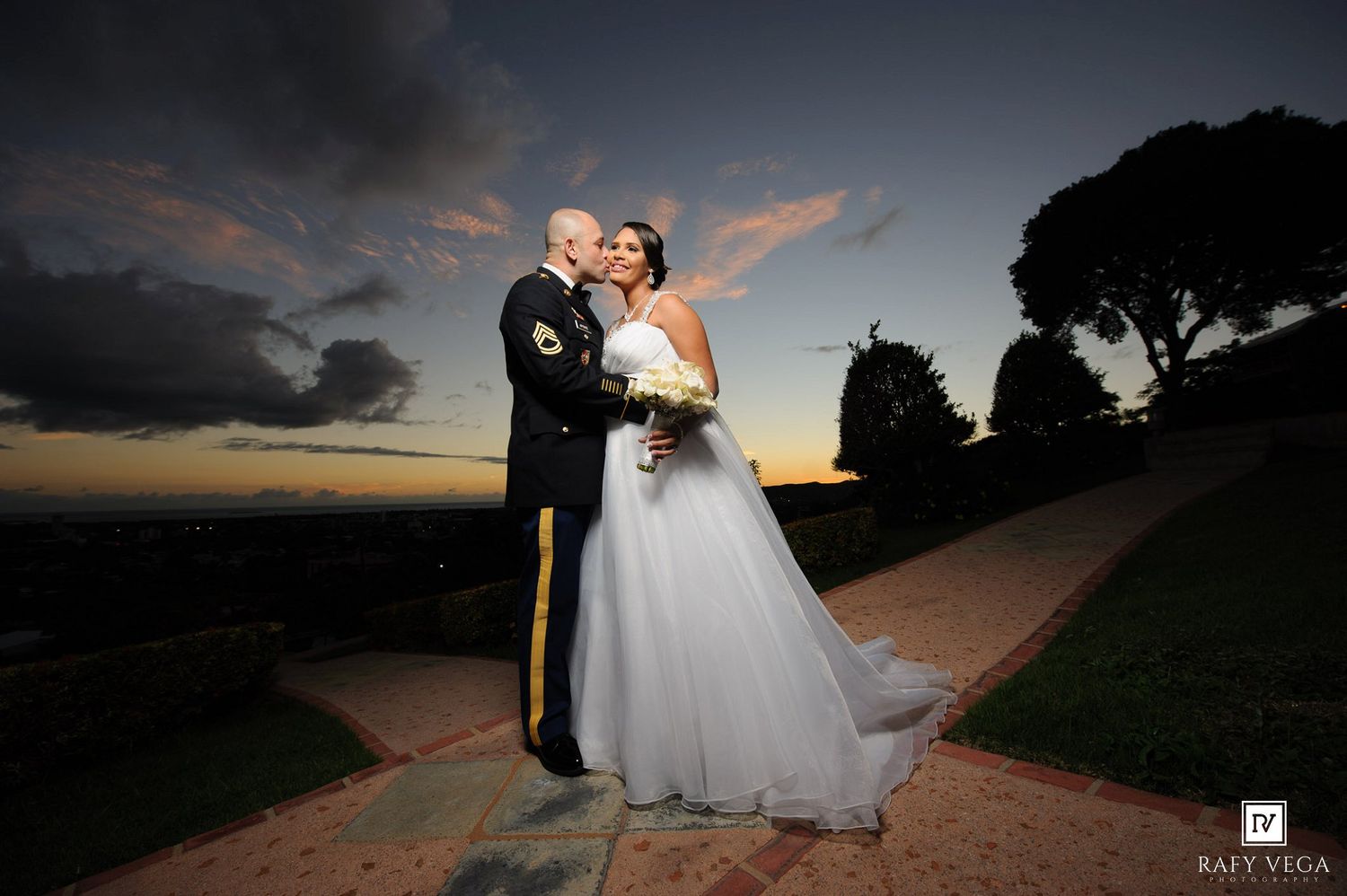 Military groom in uniform kissing bride in white gown at sunset on a scenic outdoor pathway.
