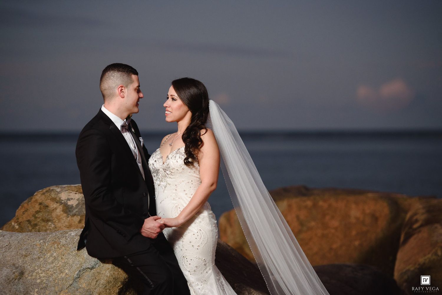 Bride and groom share a romantic moment on rocky seaside at dusk, bride wearing fitted lace gown with long veil.