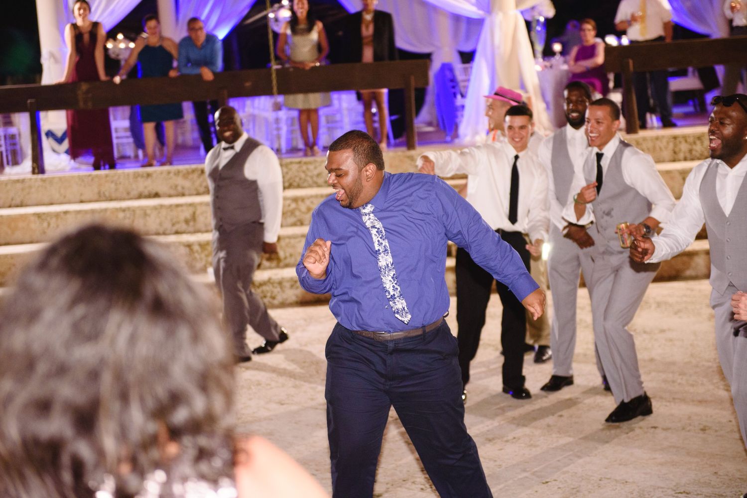 Guests dancing energetically at an outdoor wedding reception at night, with decorative purple lighting in the background.