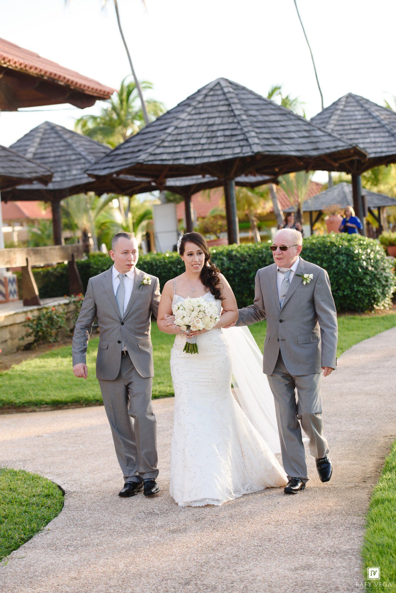 Bride in white gown walks with two men in gray suits on garden path at tropical resort wedding venue.
