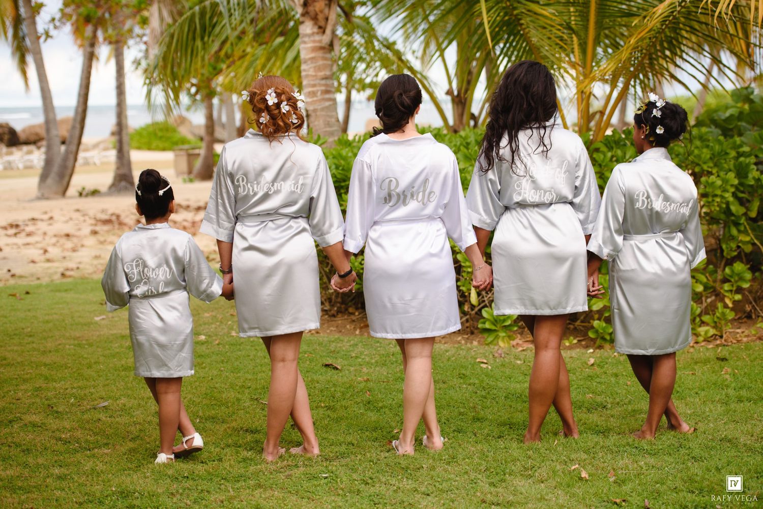 Bridesmaids and flower girl in matching silver robes holding hands on tropical beach lawn with palm trees.