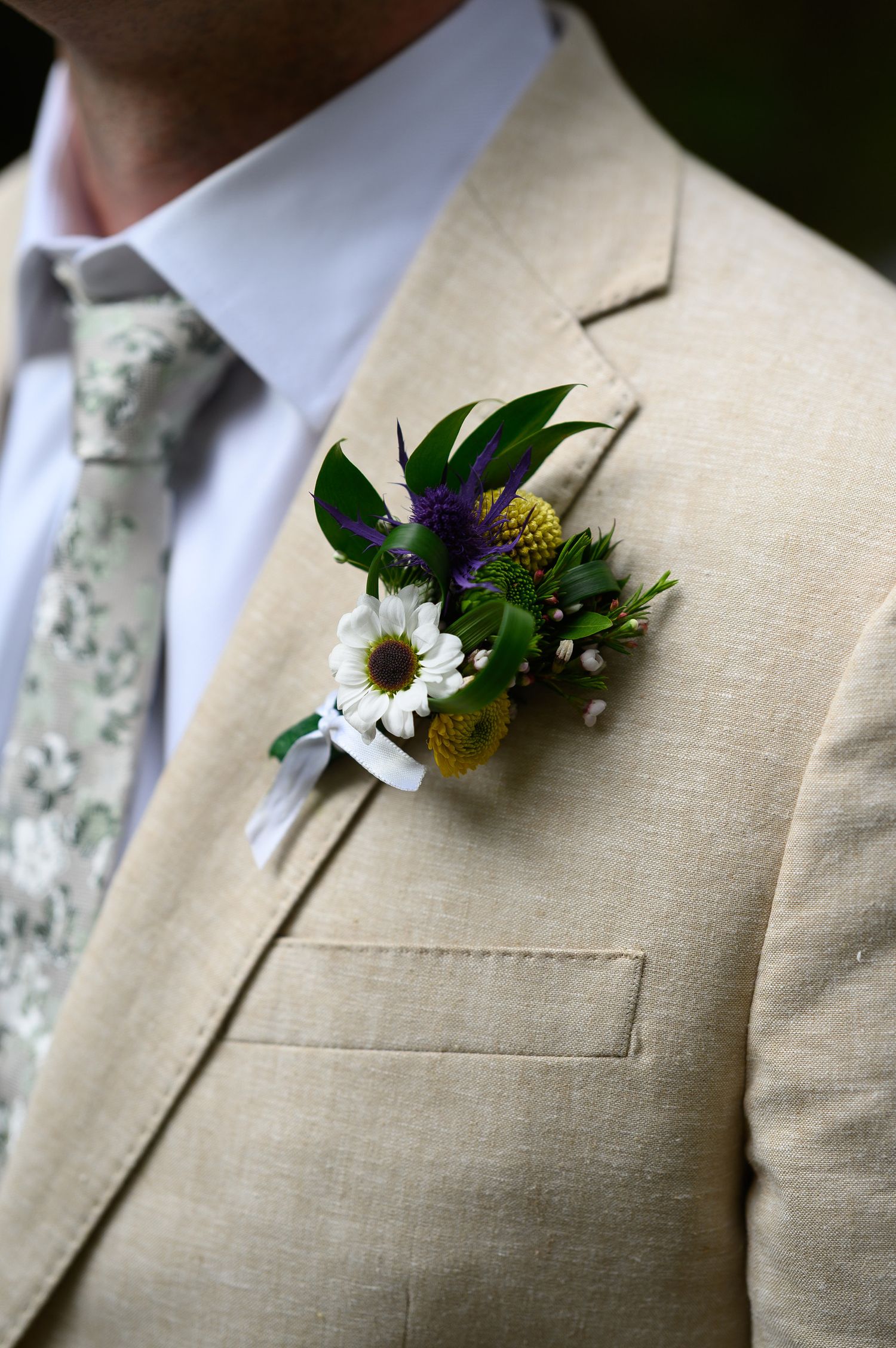 Man in tan linen suit wearing a boutonniere with white daisy, yellow craspedia, purple thistle, and greenery at a wedding.