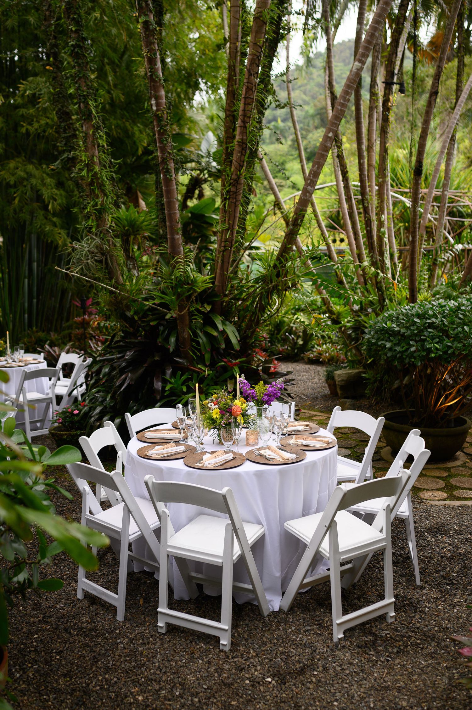 Elegant outdoor wedding reception with white round tables, chairs, and floral centerpieces set among lush tropical greenery.