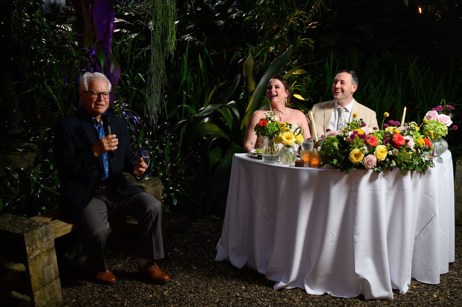 Elderly man giving a toast at an outdoor wedding reception while bride and groom smile at a flower-adorned sweetheart table.