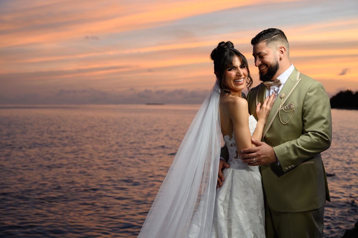 Bride and groom embrace at sunset over ocean, she in white gown with veil, he in tan suit, pink sky backdrop.