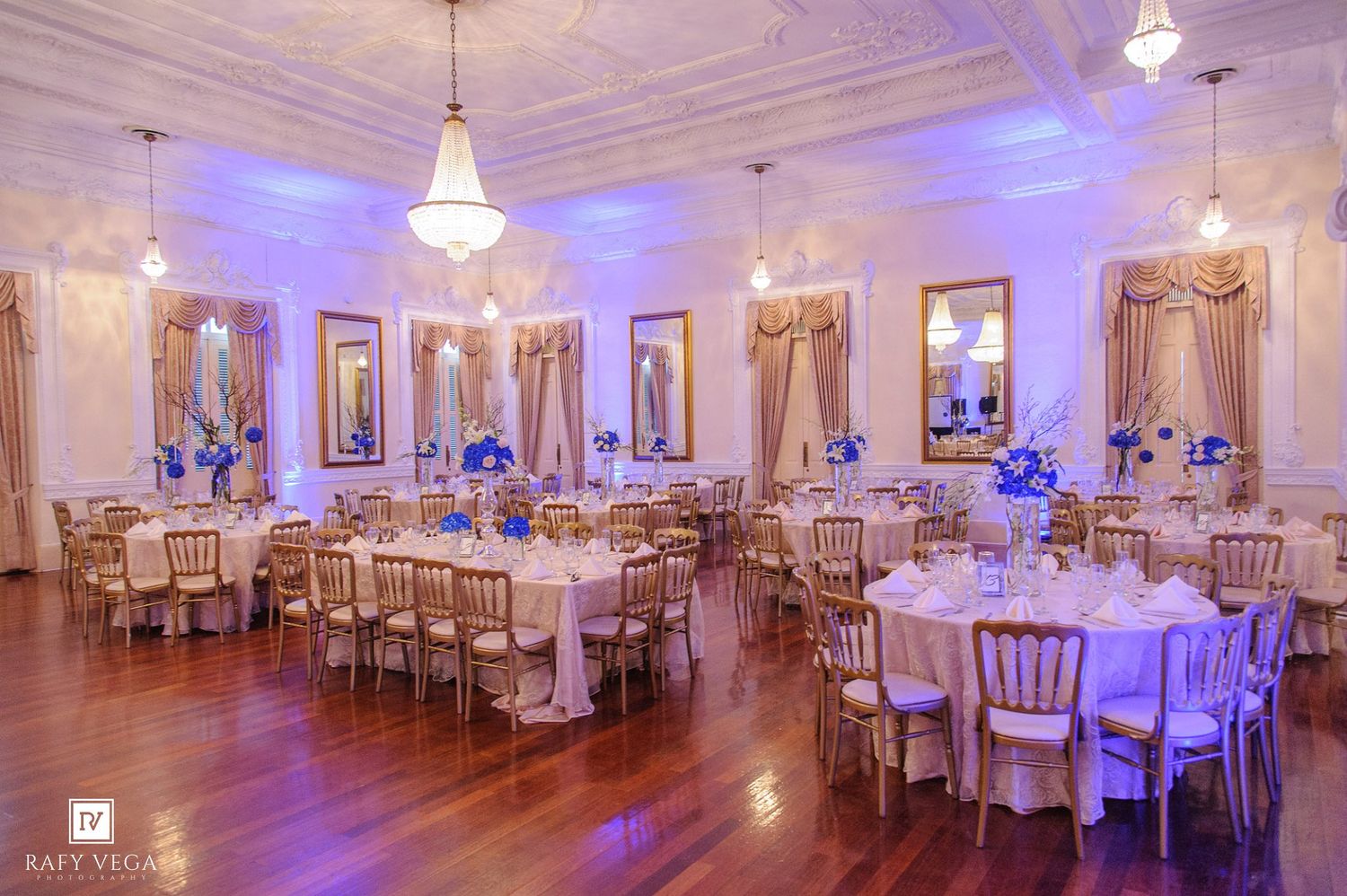 Elegant banquet hall with gold chairs, white tablecloths, blue floral centerpieces, and purple uplighting for a formal event.
