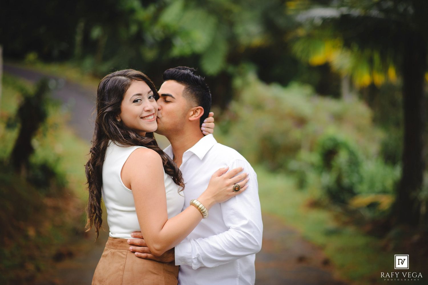 Romantic couple embracing outdoors, man kissing woman on cheek, lush tropical greenery background, engagement photo session.