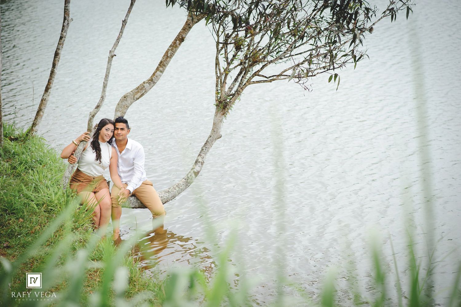 Romantic couple sitting together by a serene lake shore, leaning against a tree in a lush green outdoor setting.