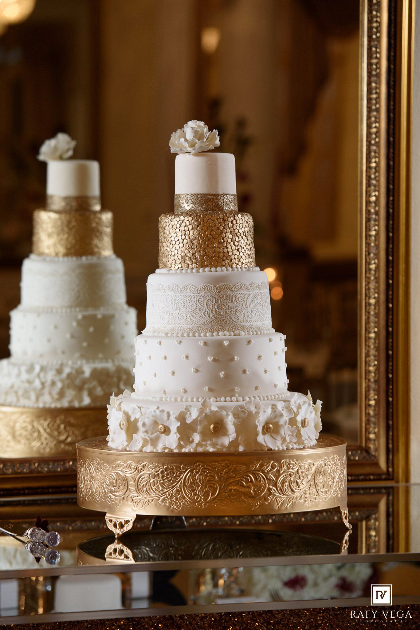 Elegant multi-tier white and gold wedding cake with floral details on a gold stand, reflected in a ornate mirror.