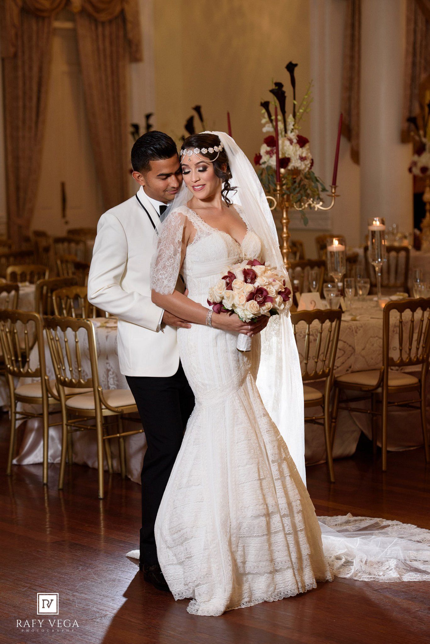 Wedding couple poses in grand ballroom, bride holding bouquet in mermaid dress with veil, groom in white tuxedo.