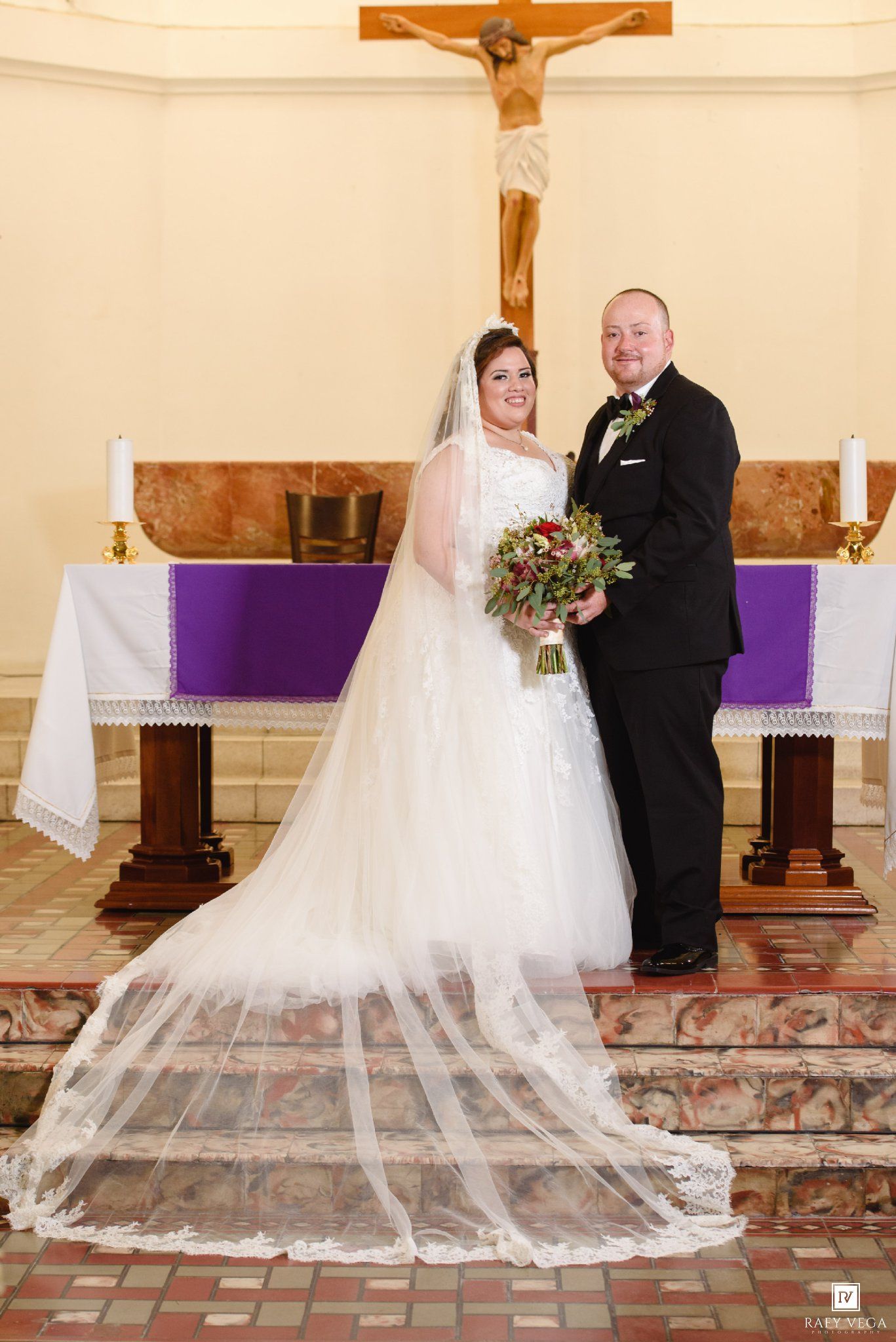 Bride and groom pose together at church altar with crucifix, bride wearing white gown with long veil holding bouquet.