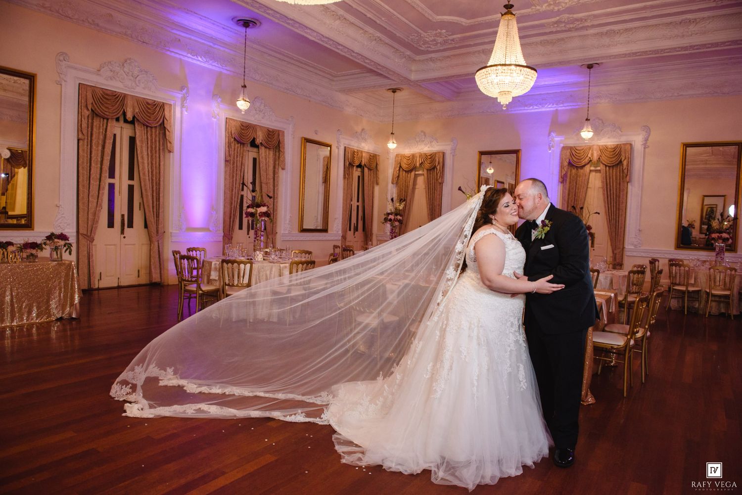 Bride and groom share a romantic first dance in an elegant ballroom, her cathedral veil flowing dramatically behind her.