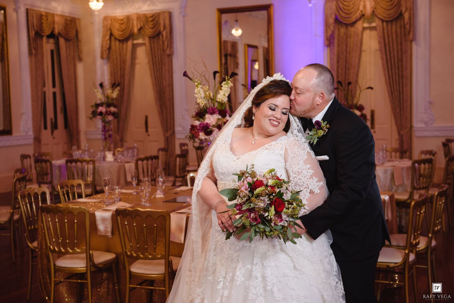 Groom kissing bride on cheek in elegant gold ballroom wedding reception, bride holding lush red and green bouquet.