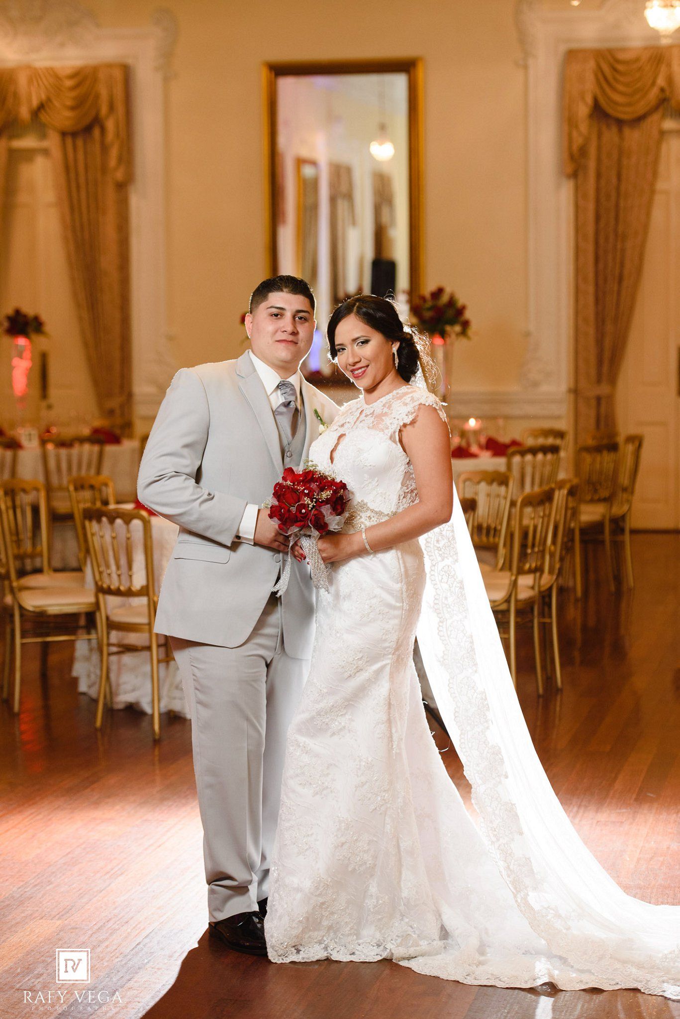 Bride in white lace gown holding red bouquet stands with groom in gray suit at elegant wedding reception venue.