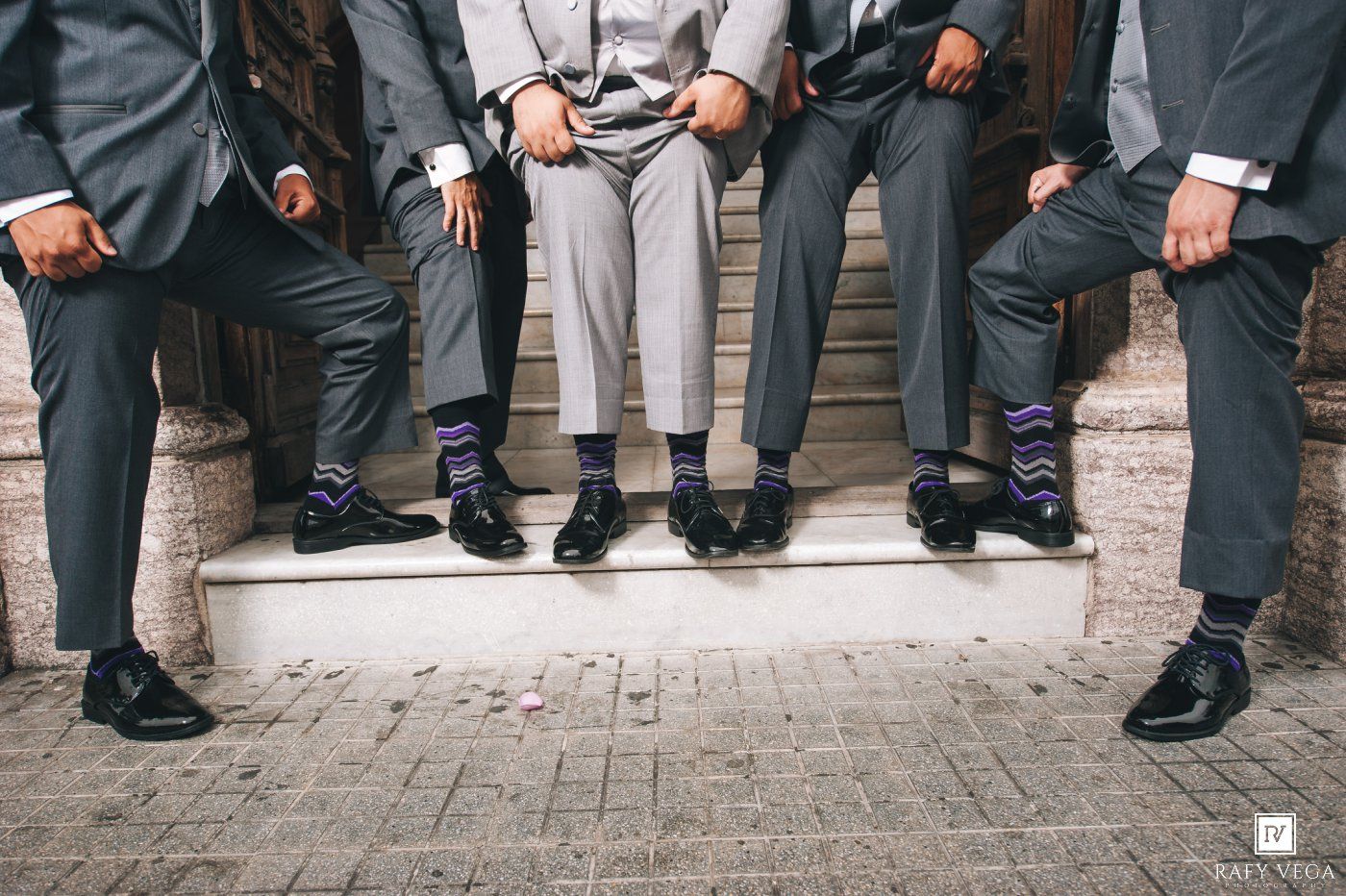 Groomsmen showing off colorful patterned socks with black dress shoes and gray suits while seated on steps.