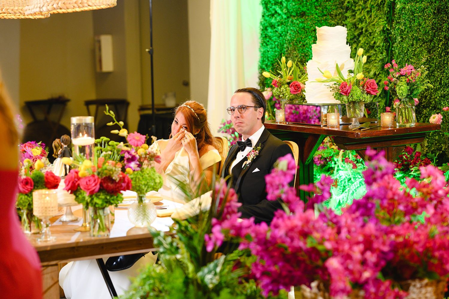 Bride and groom seated at floral-decorated wedding reception table with pink blooms and green hedge backdrop.