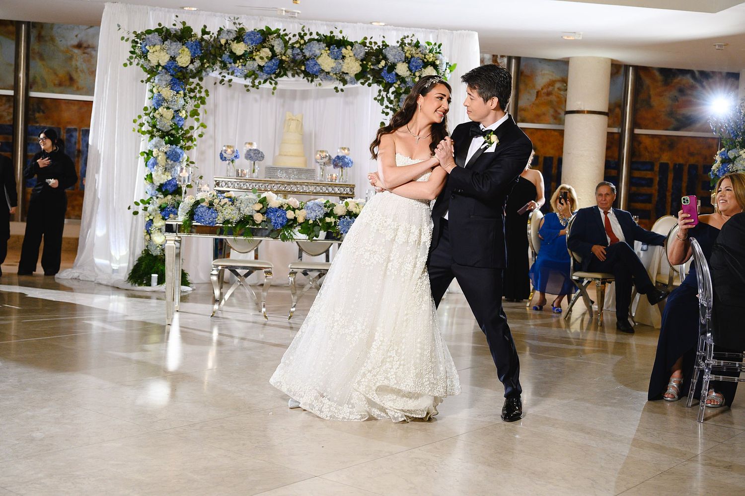 Bride and groom share first dance at wedding reception with blue and white floral arch.