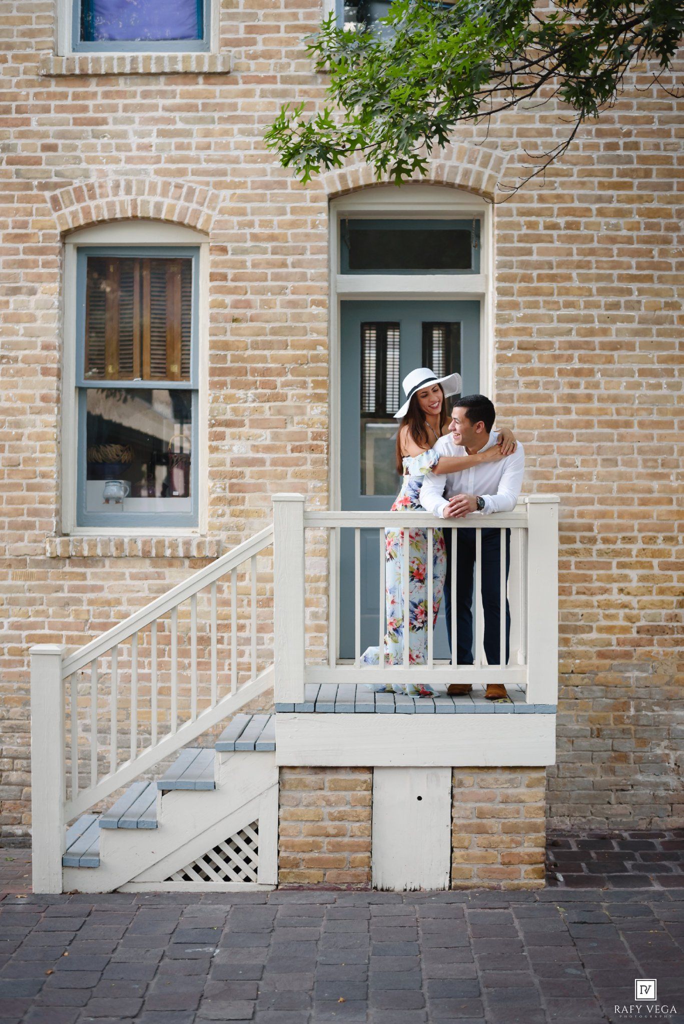 Couple embracing on a charming brick building porch, woman in floral jumpsuit and white hat.