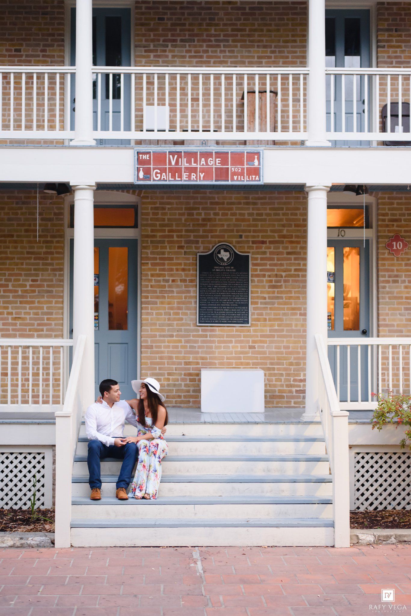Couple sitting on steps of The Village Gallery brick building, woman in floral dress and hat, man in white shirt.