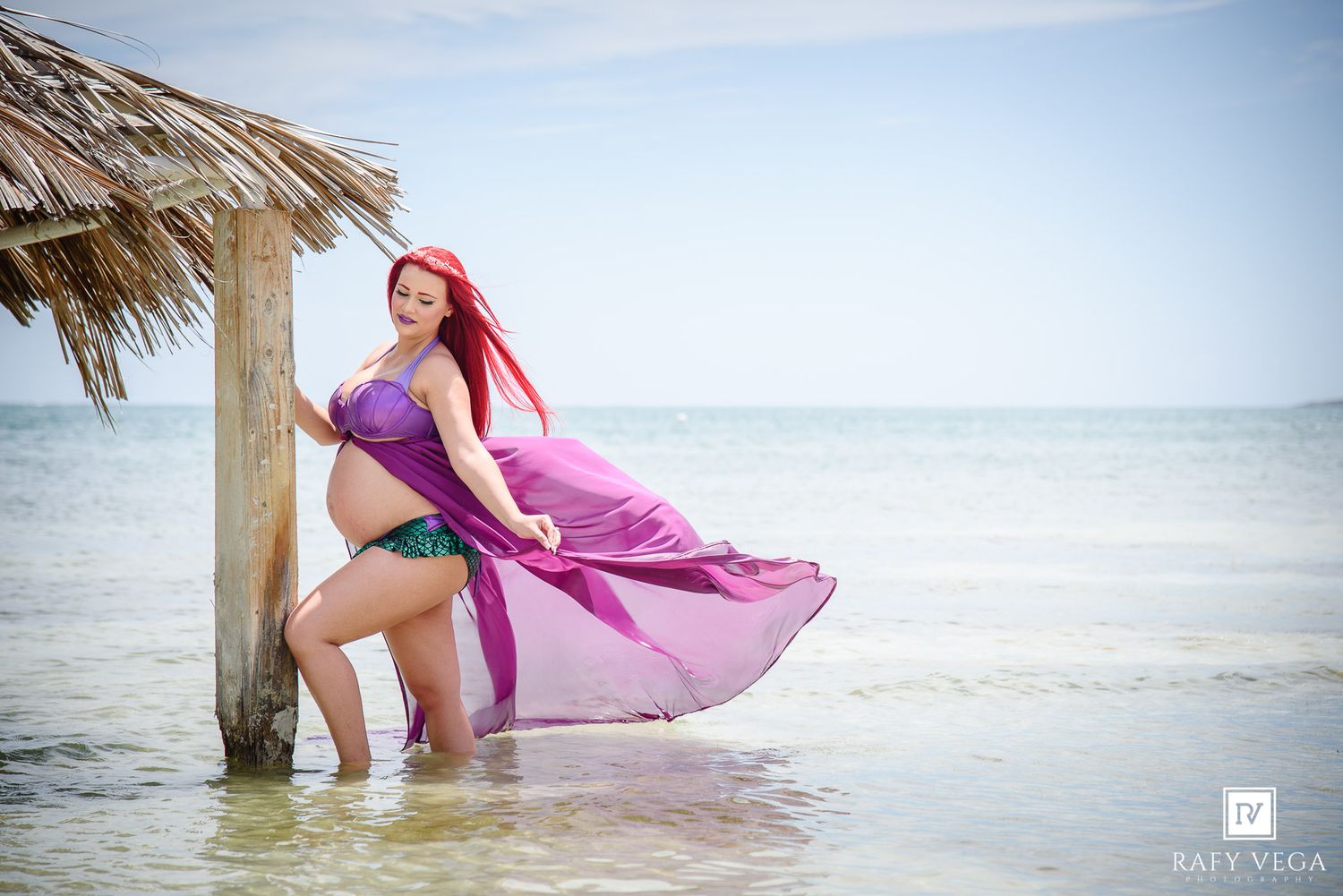 Pregnant woman in purple maternity gown standing in ocean water beside a thatched hut post on a tropical beach.