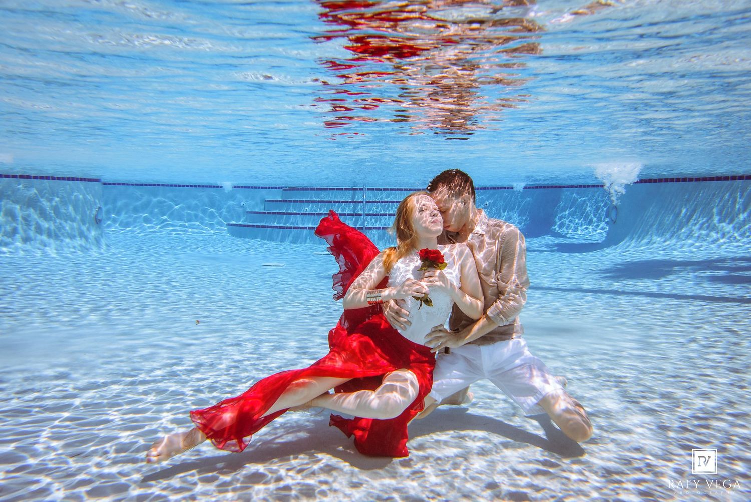 Couple in elegant red and white outfits pose romantically in an underwater photoshoot in a clear blue pool.