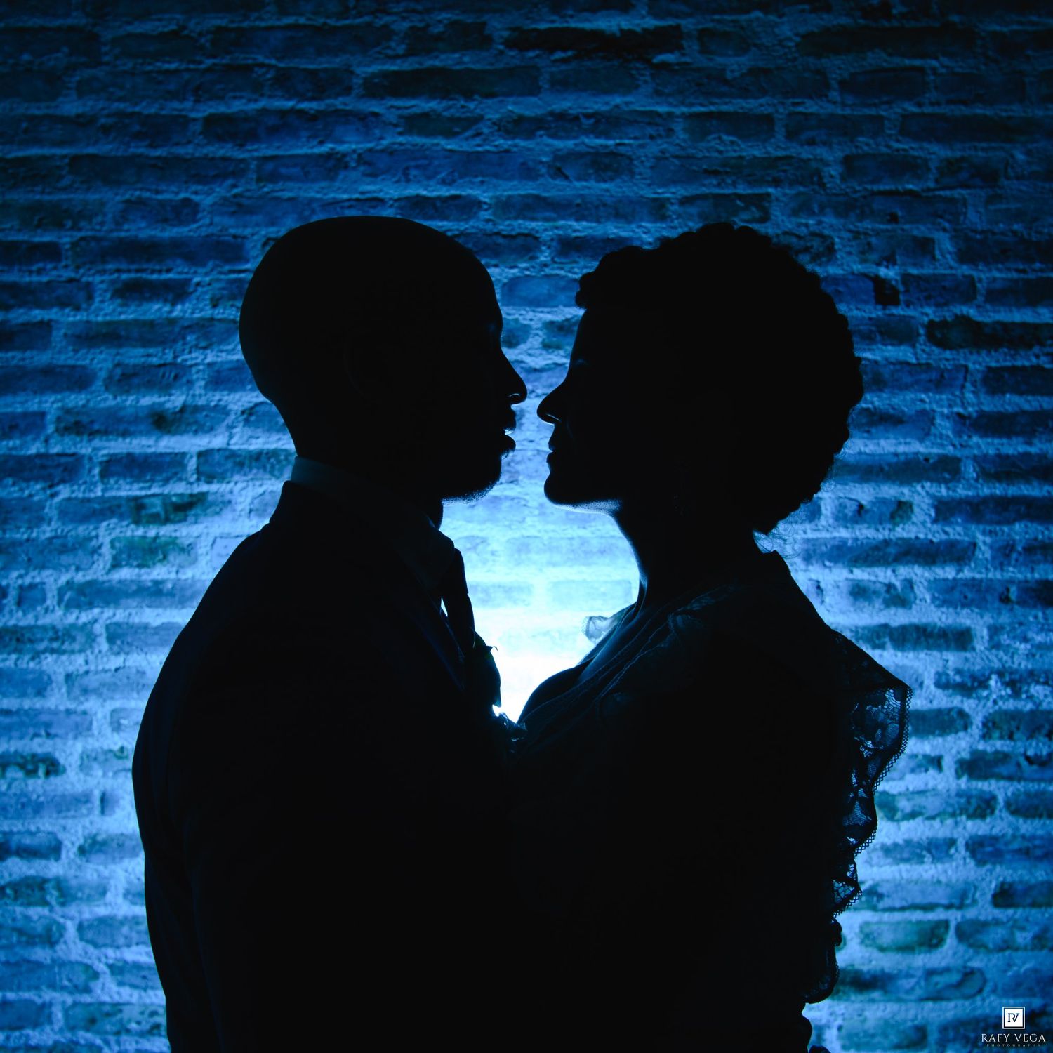 Romantic silhouette of a couple facing each other against a blue-lit brick wall backdrop.