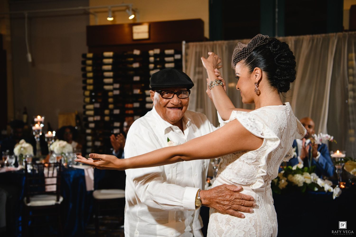 Couple dancing joyfully at wedding reception, man in white shirt and cap, woman in lace dress, elegant venue.