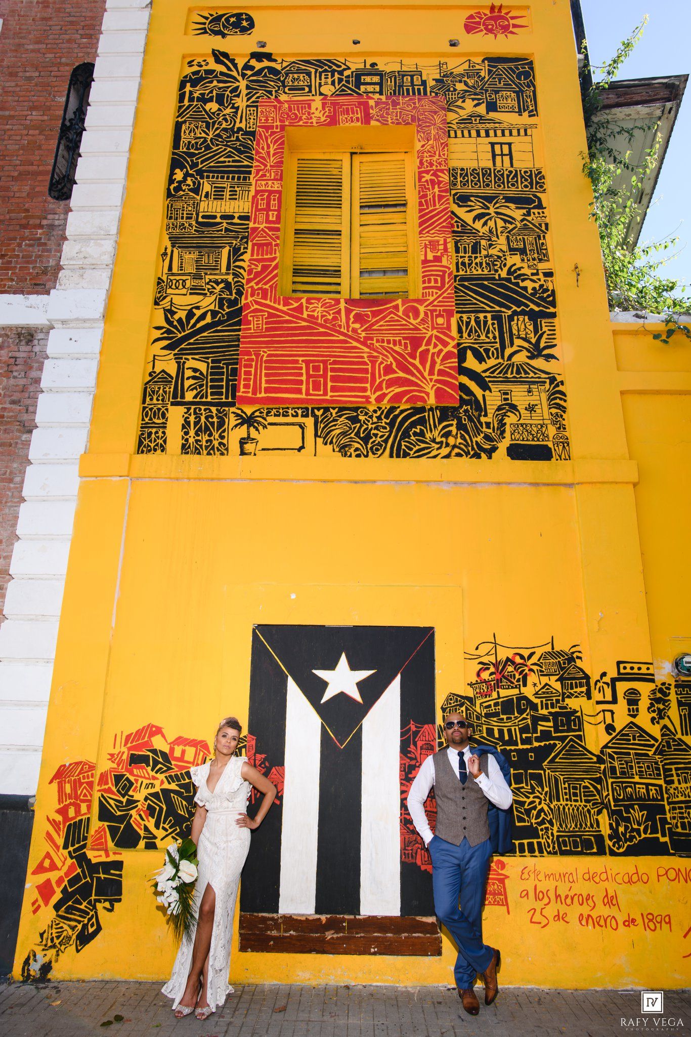 Couple poses before vibrant yellow mural with Puerto Rican flag in Old San Juan, bride in white gown, groom in blue suit.