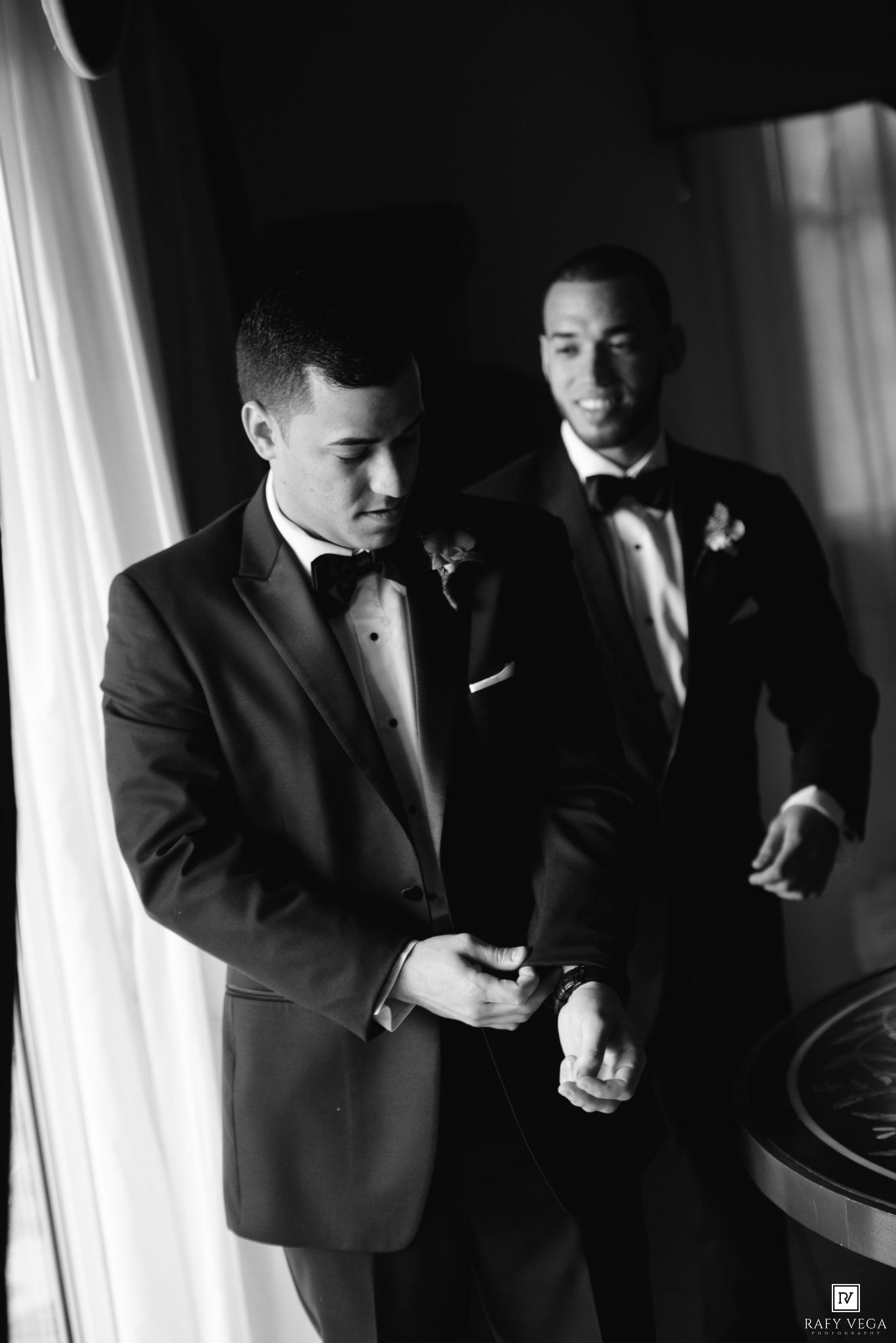 Two grooms in black tuxedos with bow ties and boutonnieres prepare together in a black and white wedding photo.