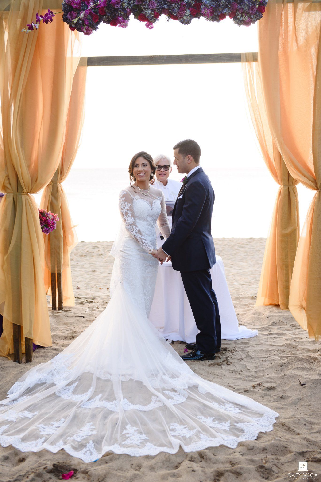 Bride in lace mermaid gown holds hands with groom under golden draped arch during romantic beach wedding ceremony.