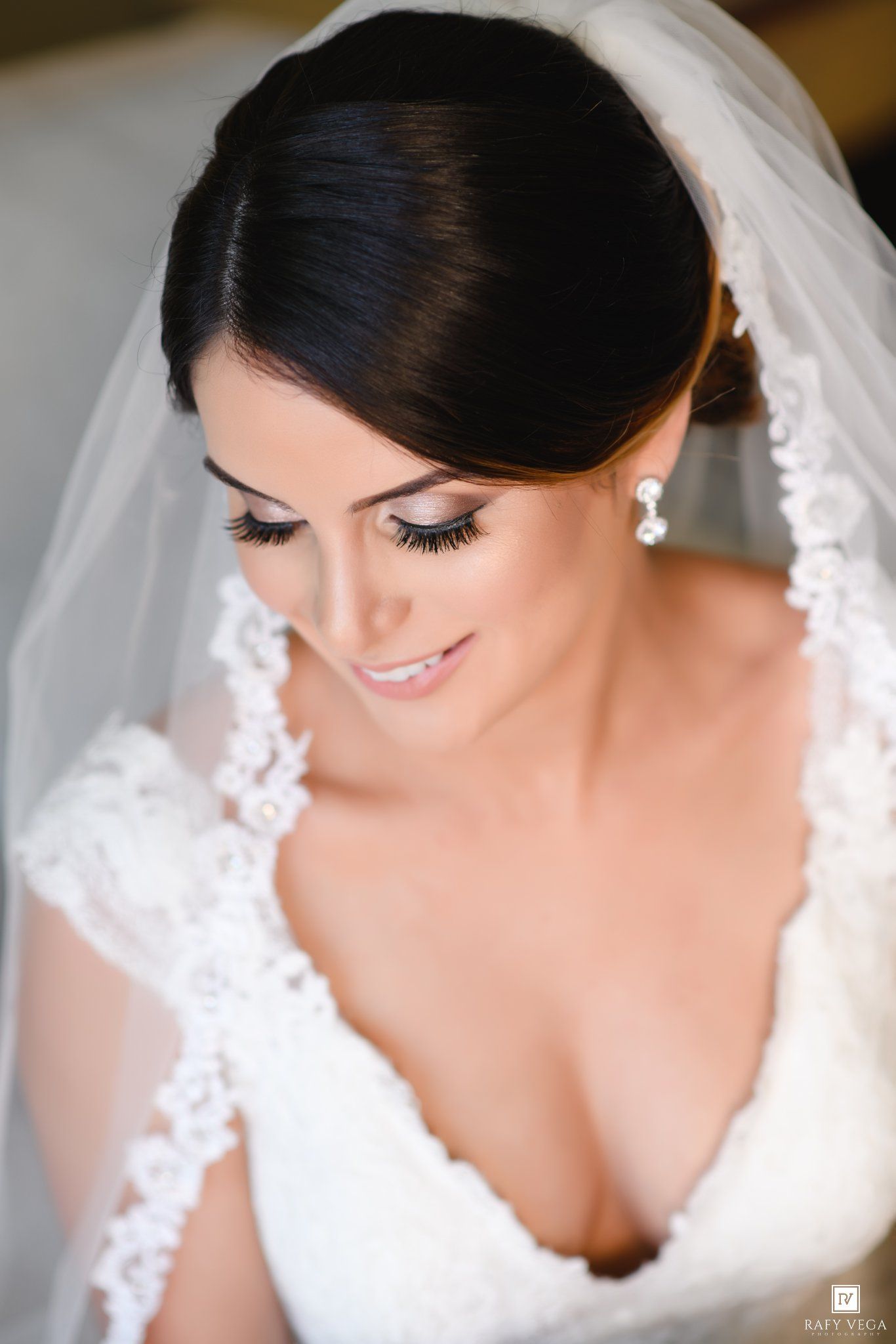 Elegant bride with dark updo hair, lace veil, pearl earrings, and white gown, wearing natural bridal makeup.