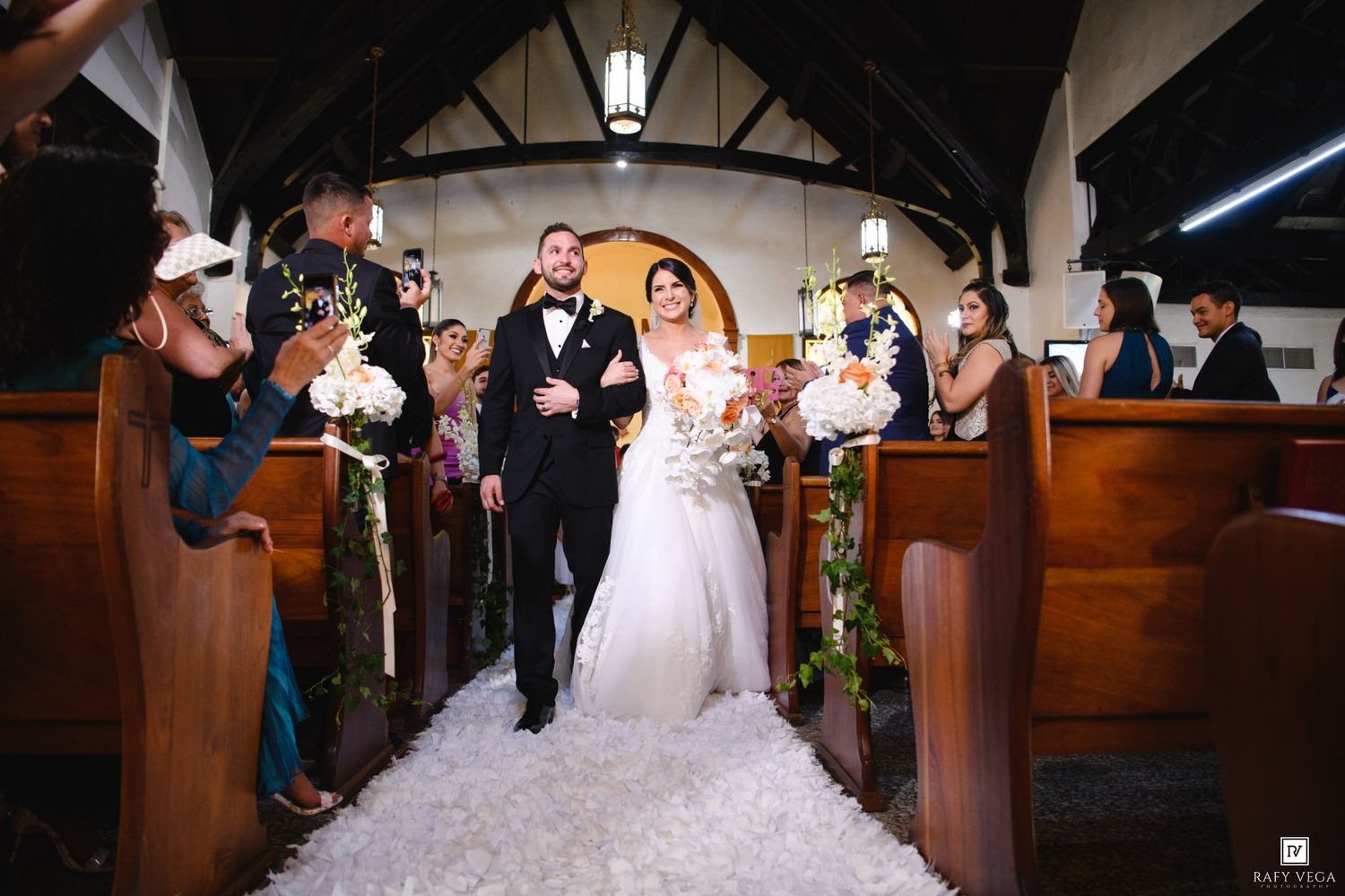 Newlywed couple walks down church aisle in wedding ceremony, bride in white gown holding bouquet, guests celebrating.