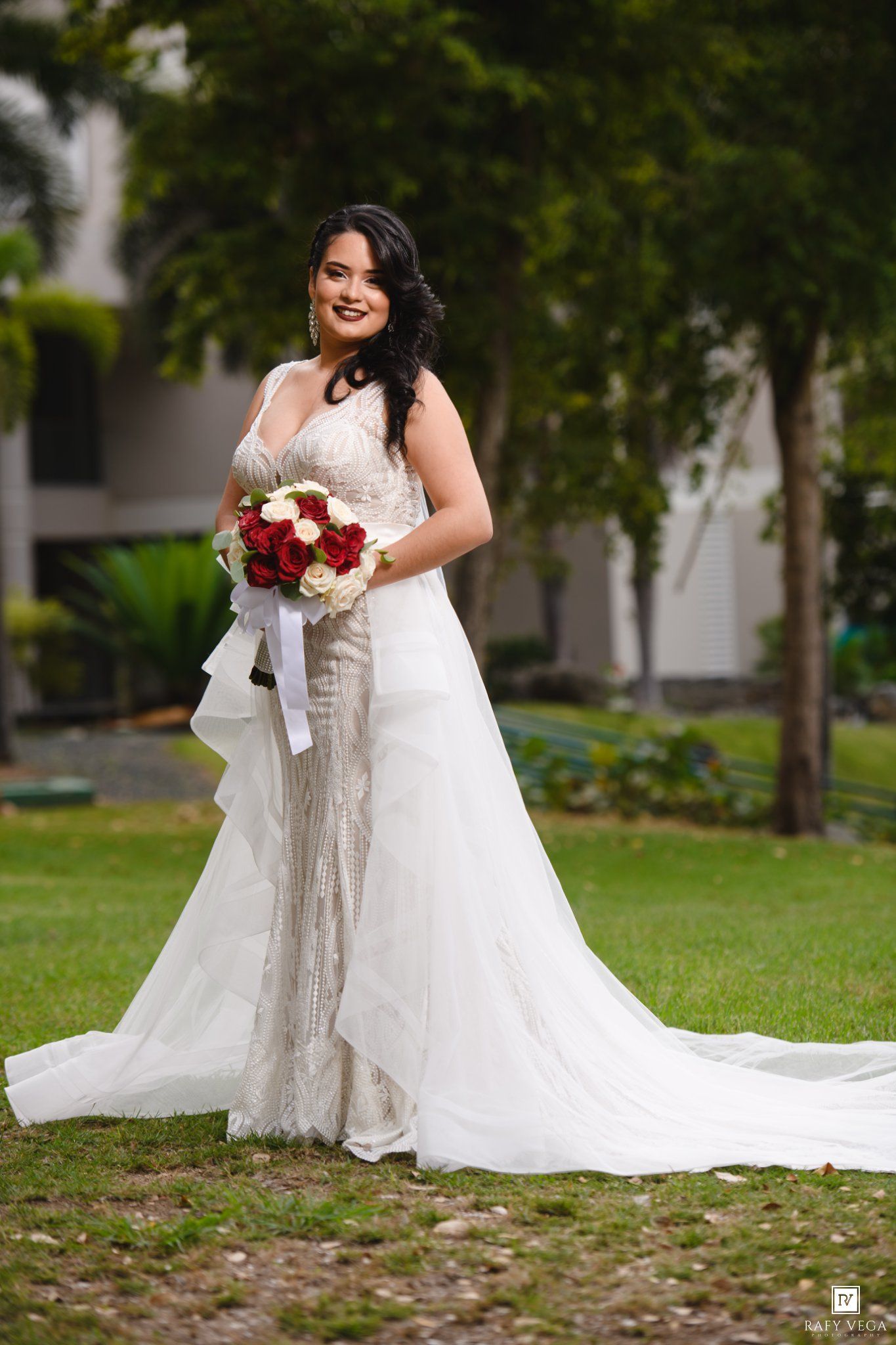 Bride in white lace mermaid gown holding red and white rose bouquet, posing outdoors on green lawn with trees.