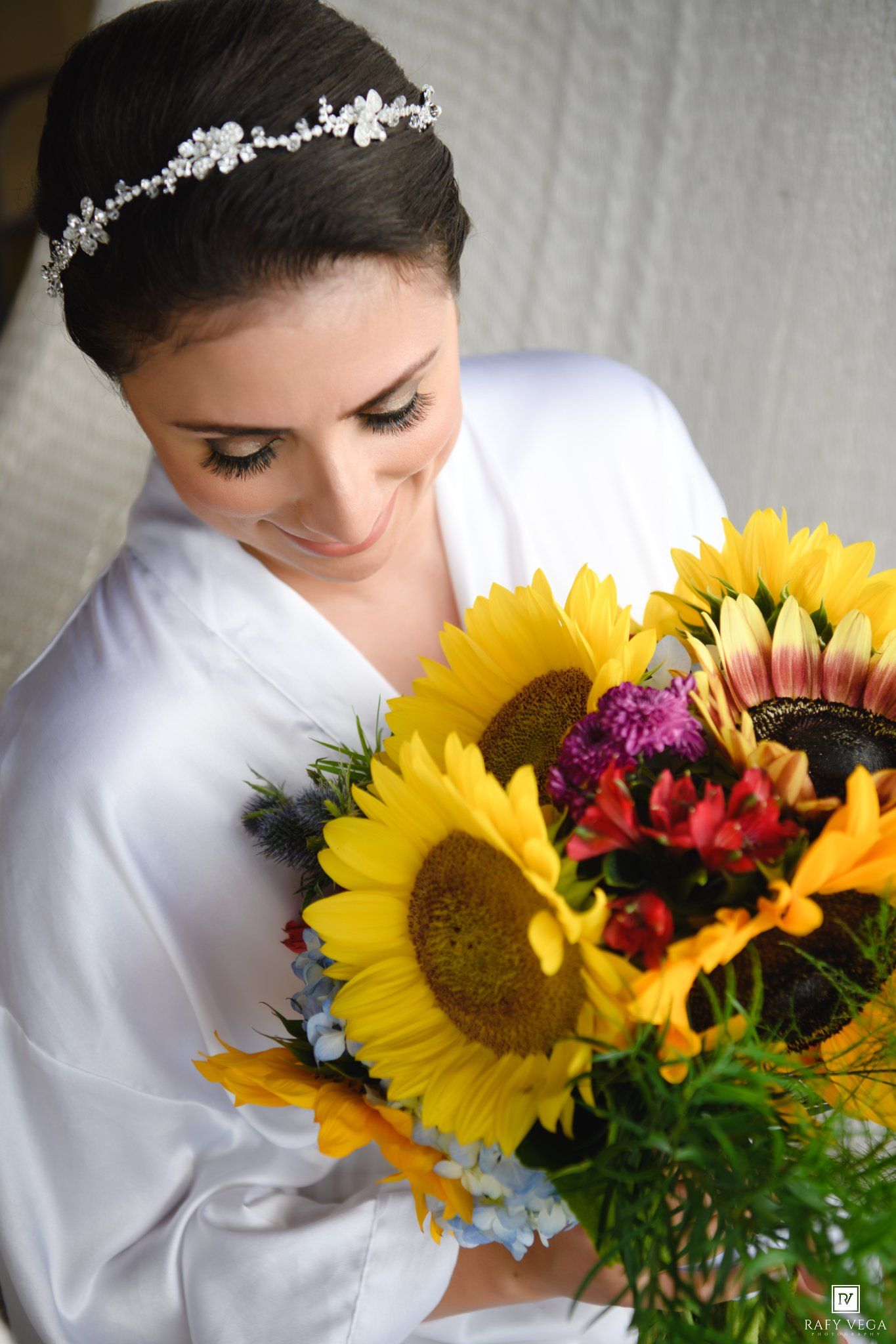 Bride in white robe holding vibrant sunflower bouquet with colorful blooms, wearing crystal hair vine.