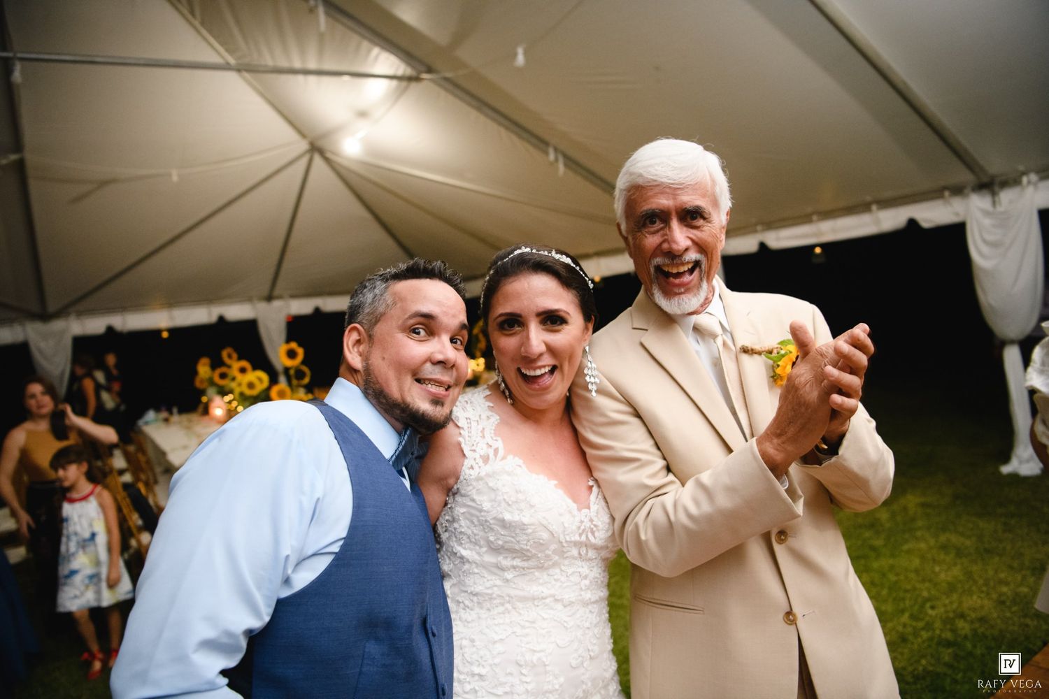 Bride and groom celebrate with older gentleman at outdoor wedding reception under white tent.