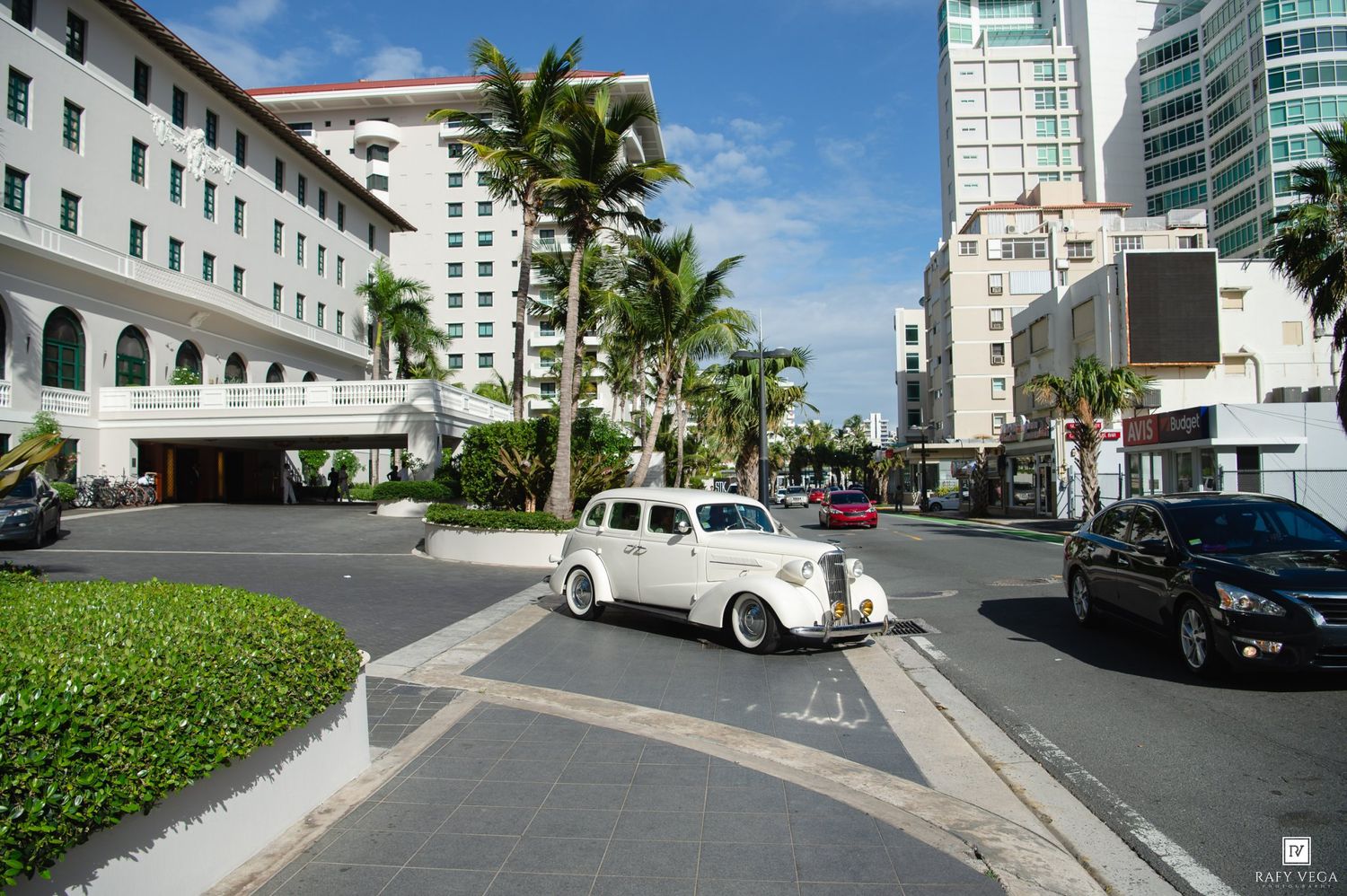 Vintage white classic car driving on a palm-lined street with luxury hotels in a tropical city.