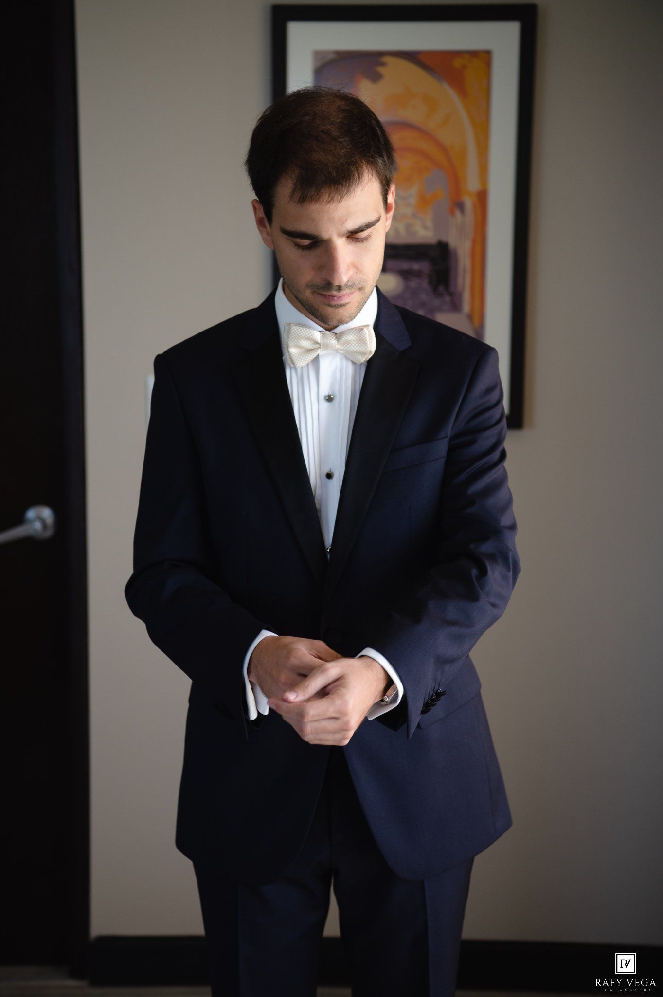 Elegant groom in navy tuxedo with ivory bow tie adjusting cufflinks before wedding ceremony.