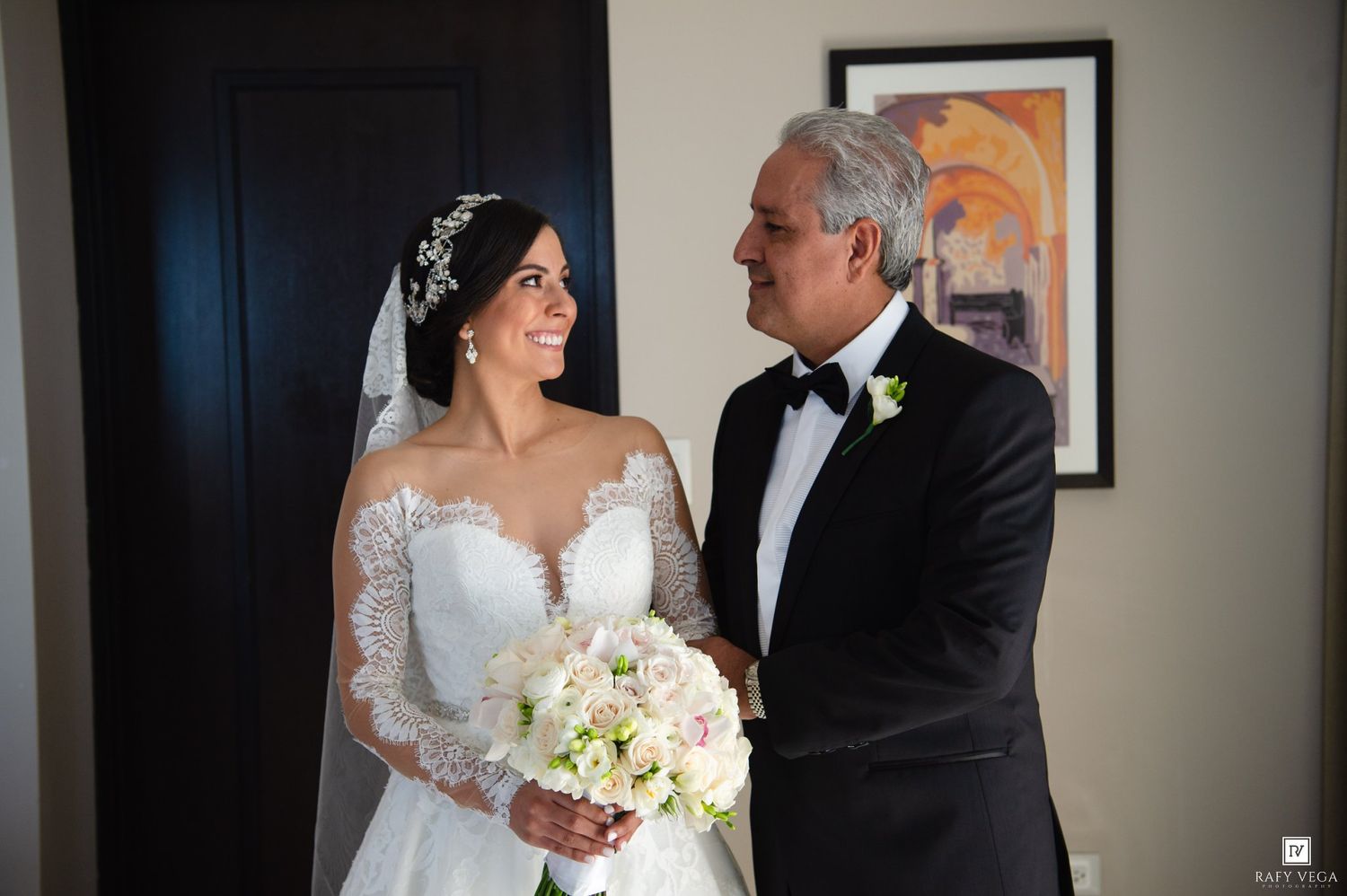 Bride in lace wedding gown with veil and bouquet shares a tender moment with older man in tuxedo indoors.