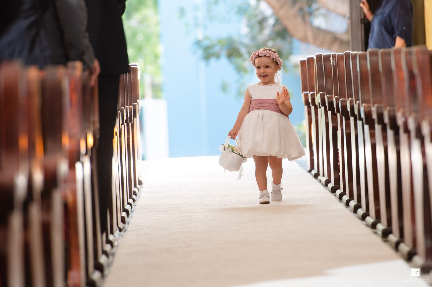 Adorable flower girl in white dress walks down wedding aisle carrying basket, wearing floral crown.