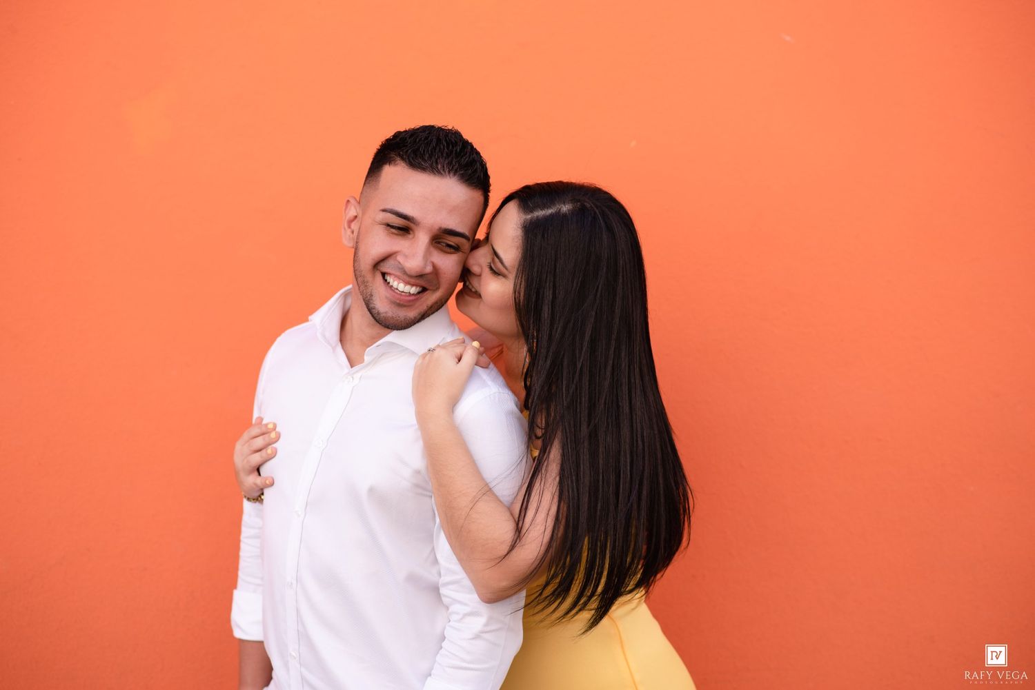 A joyful couple embracing against a vibrant orange wall, woman kissing man's cheek while he smiles broadly.