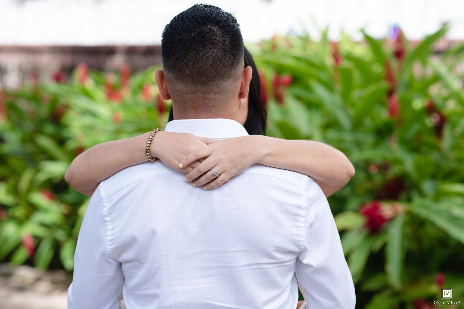 Couple embracing outdoors, woman's hand with ring on man's shoulder in white shirt, tropical garden background.