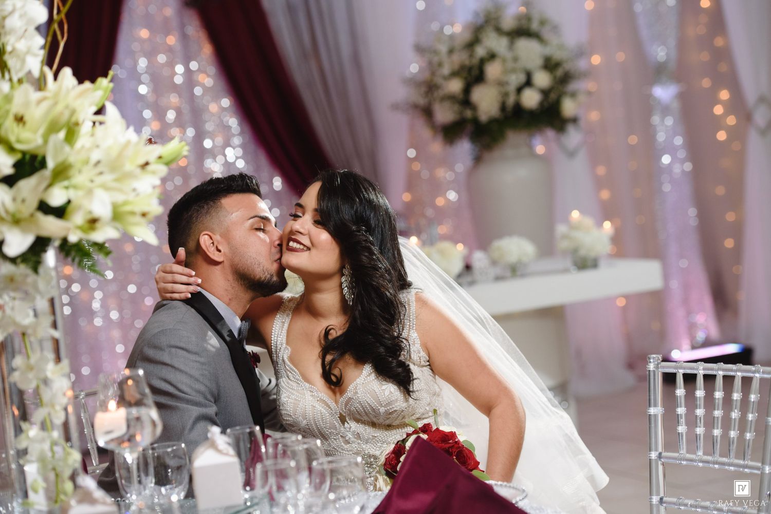 Bride and groom share a romantic kiss at elegantly decorated wedding reception with white floral arrangements and fairy lights.