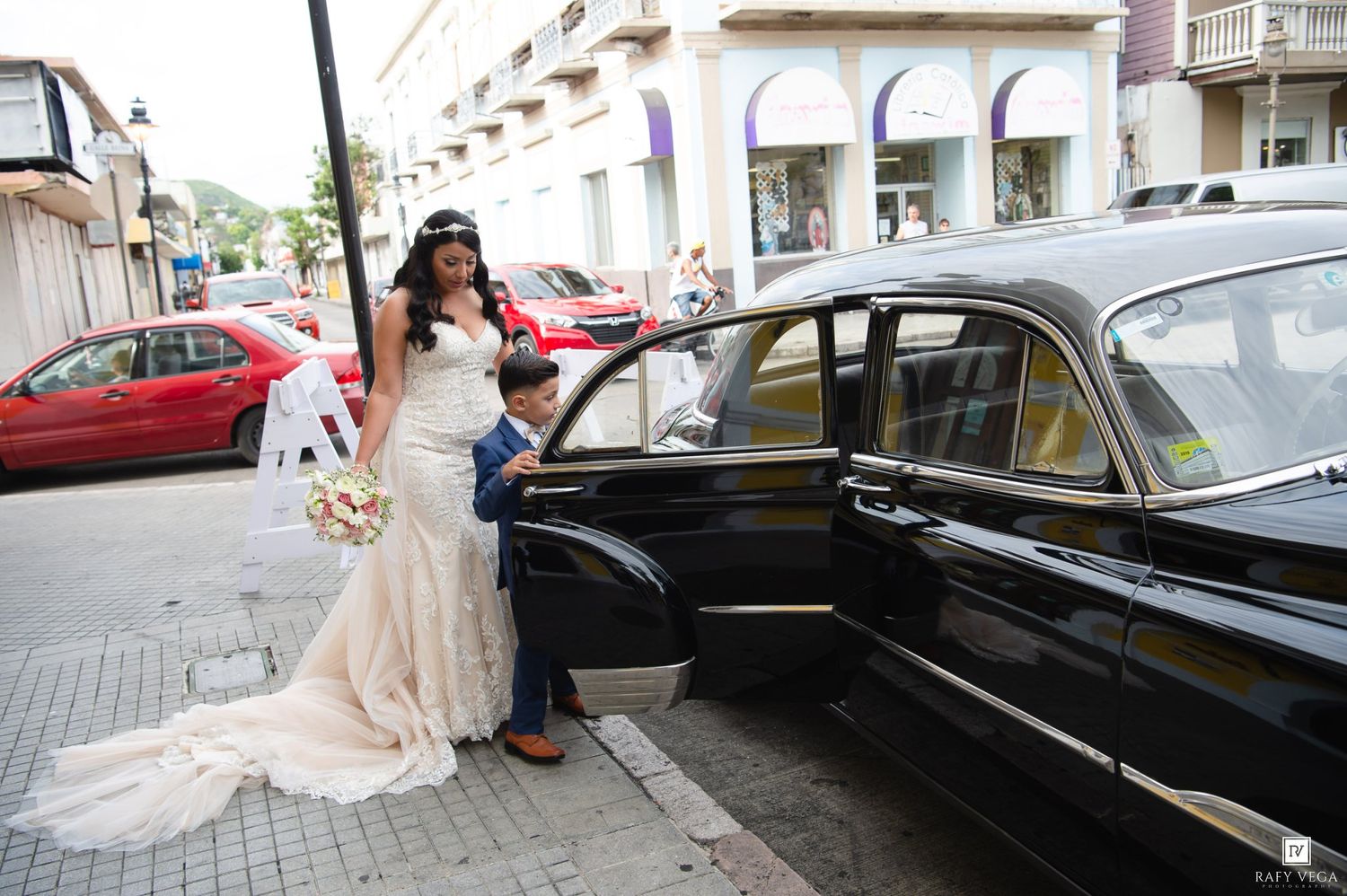 Bride in white wedding gown with a young boy opening the door of a classic black vintage car on a city street.