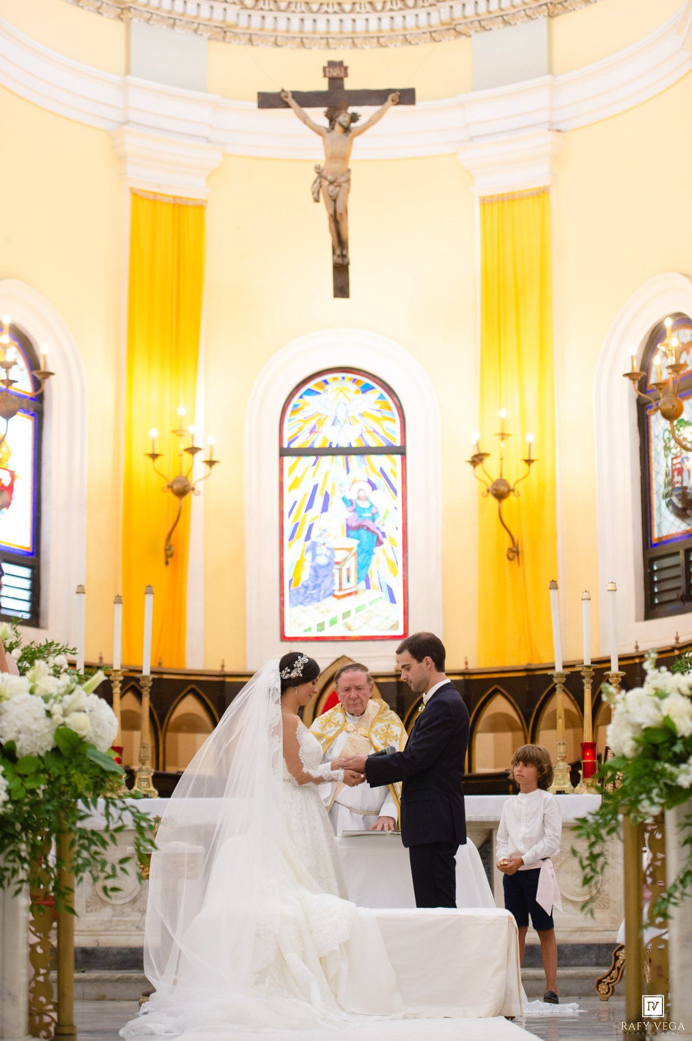 Bride and groom exchange vows at a Catholic church altar adorned with white floral arrangements and stained glass windows.