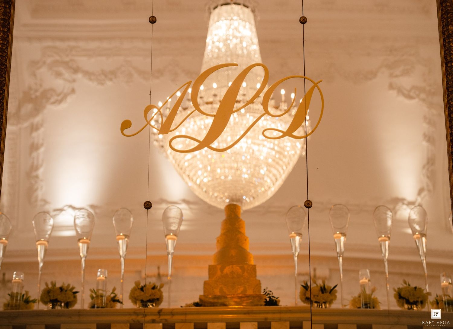 Elegant wedding reception backdrop with gold monogram ALD, crystal chandelier, tiered cake, and candlelit table display.