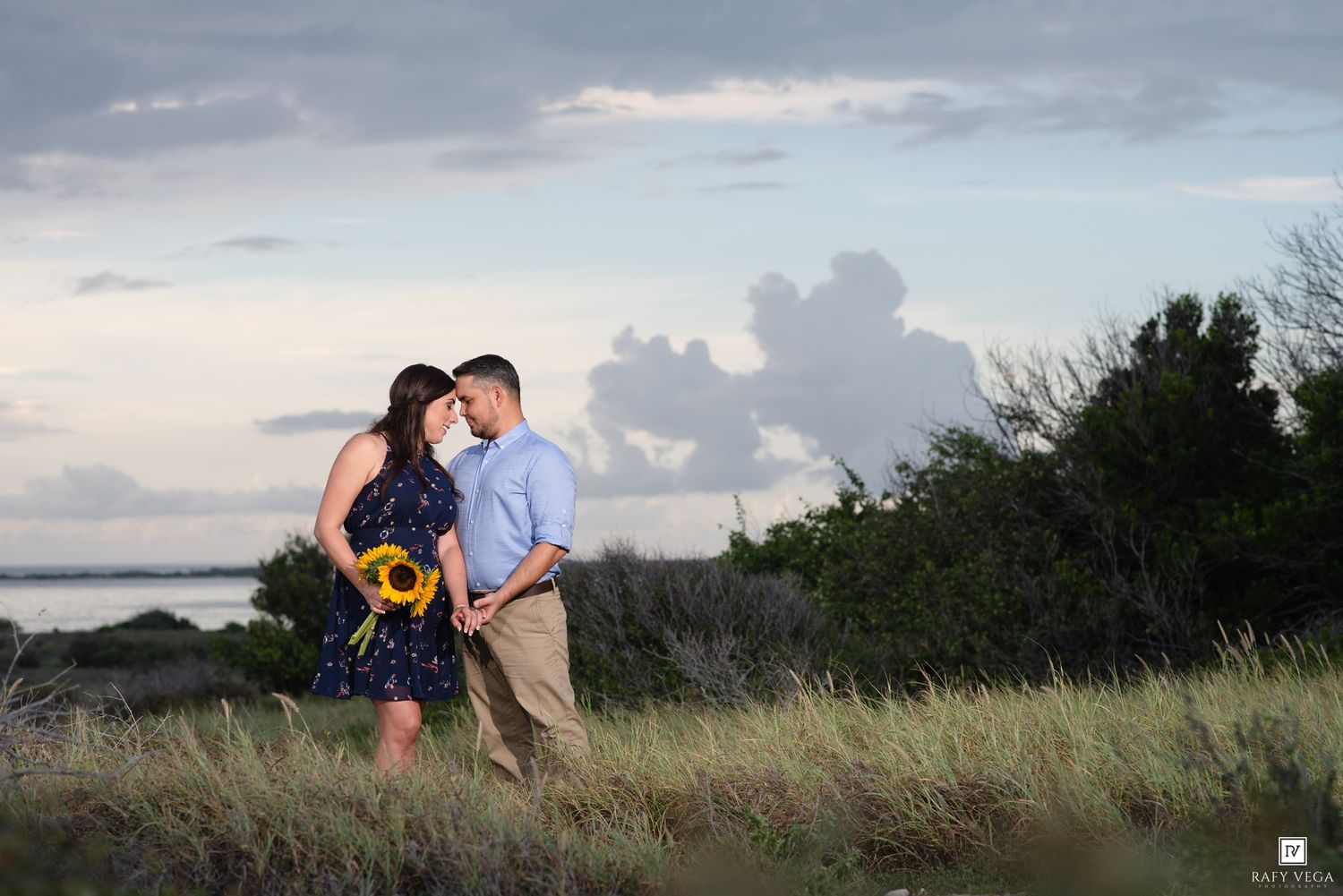 Couple embracing in a grassy field at sunset, woman holding sunflowers, with a cloudy sky and water in the background.