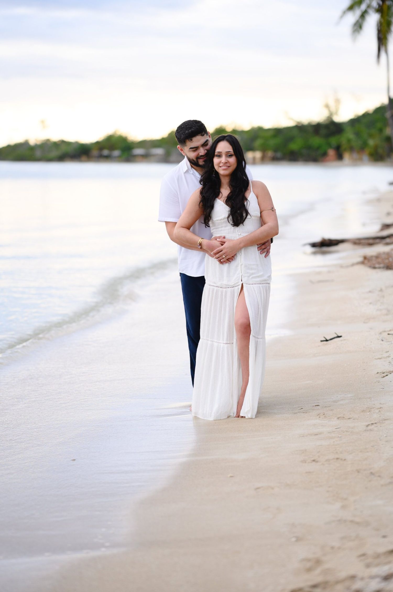 Couple embracing on a sandy beach at sunset, woman in white maxi dress, man in white shirt and navy pants.