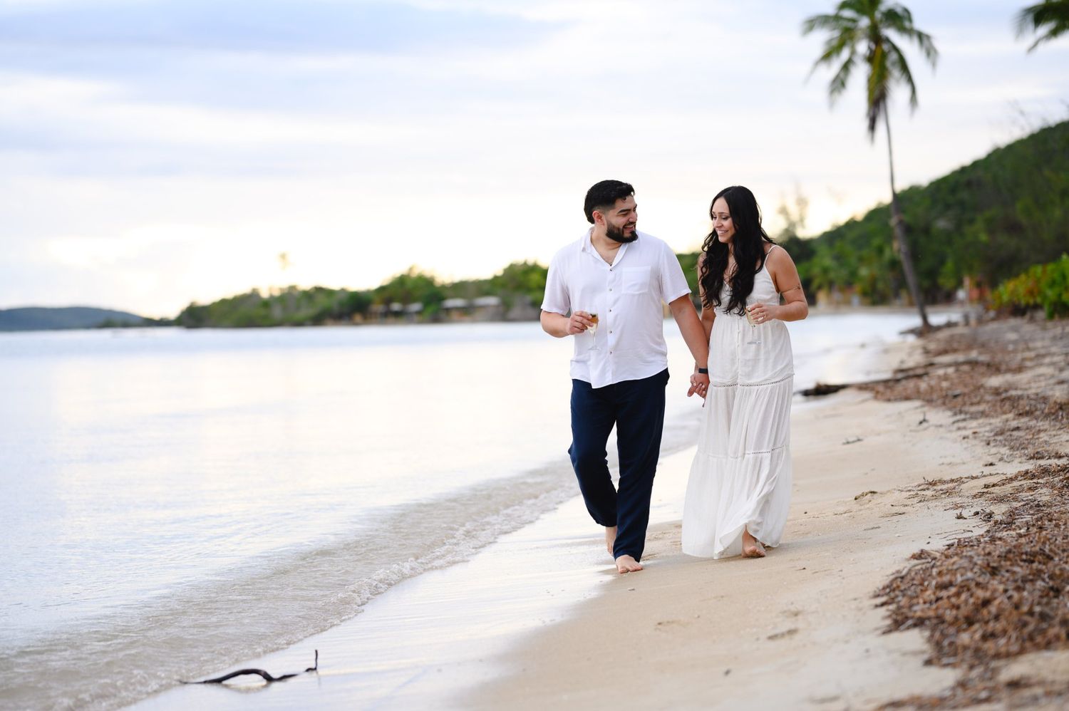Couple walking hand-in-hand on a tropical beach at sunset, wearing white outfits against a serene ocean backdrop.
