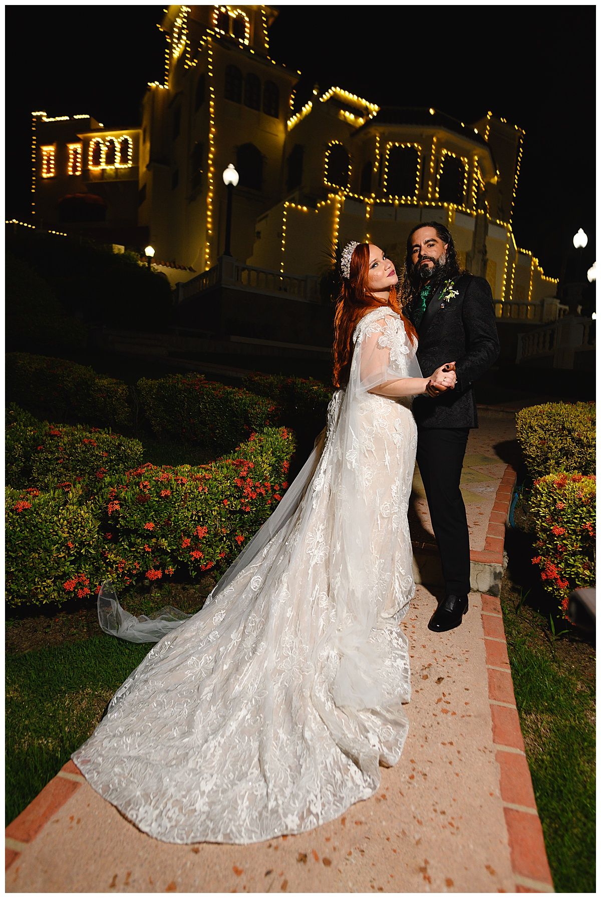 Bride in lace gown with veil and groom in black suit pose at night outside a lit historic building.