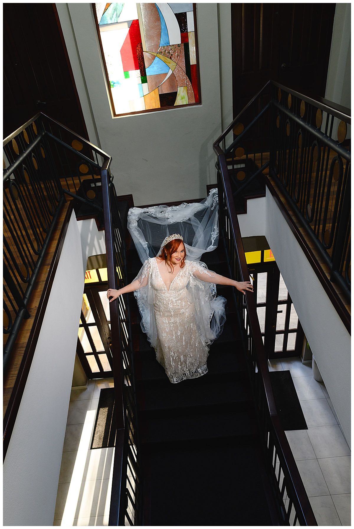 Bride in white gown and veil descends grand staircase beneath colorful stained glass window.